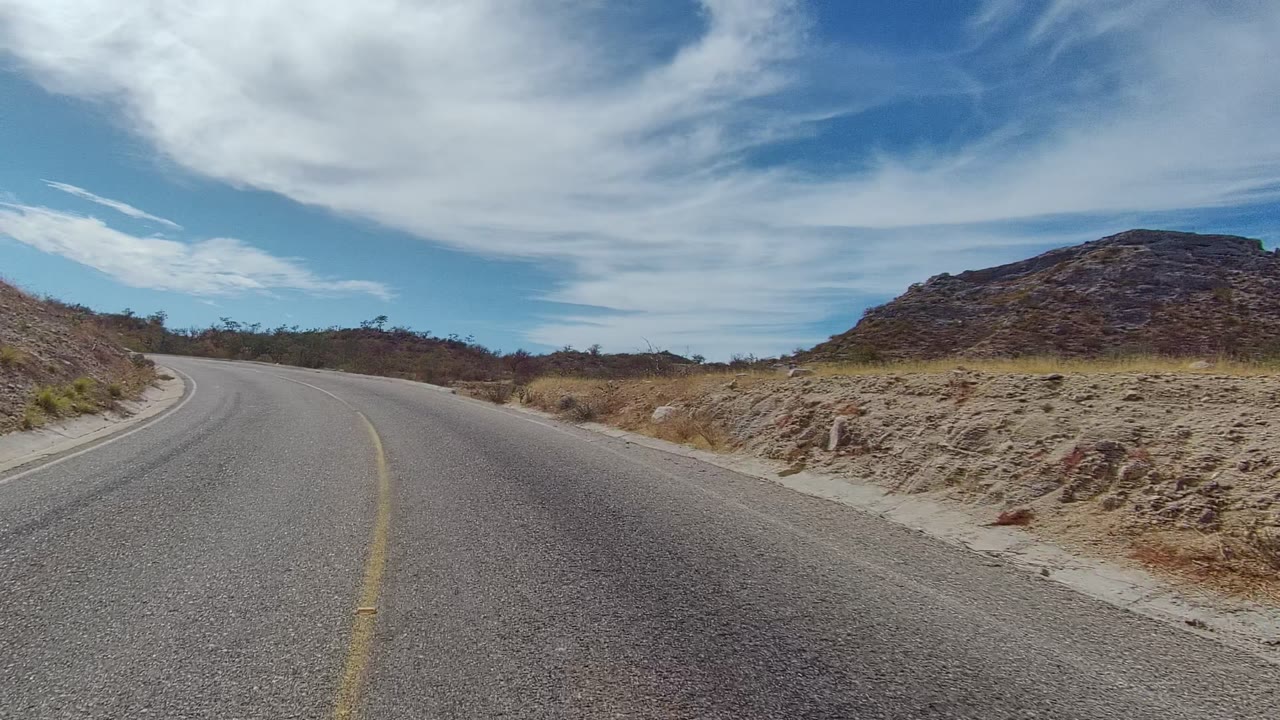 A wider view of the East Cape Los Cabos road shows the curve opening ahead, with scrub-covered shoulders, low hills, a blue sky with sweeping white clouds, and no visible people.