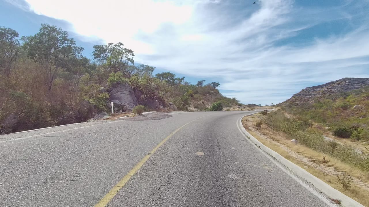 Road-level view in East Cape Los Cabos, Mexico, showing a curving asphalt lane with a yellow center line, dry brush, rocky slope, blue sky, and no visible people.