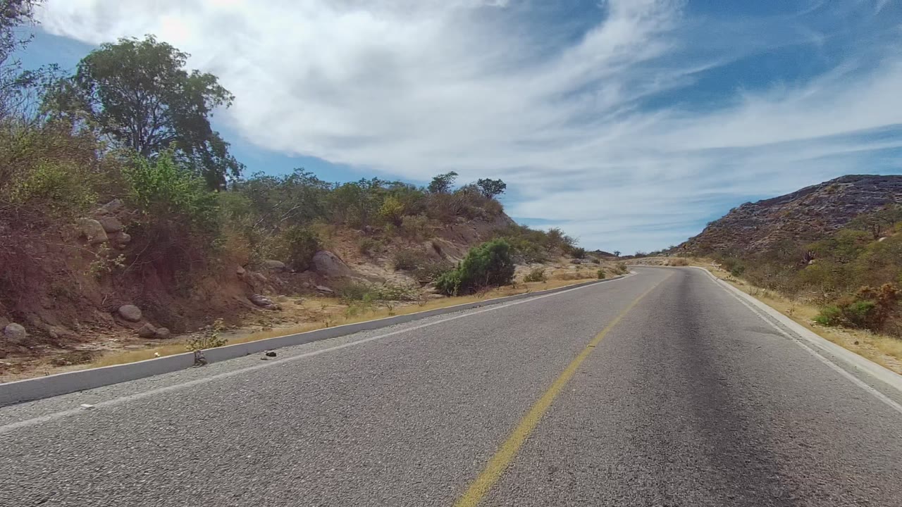 Paved road in East Cape Los Cabos, Mexico, running toward a low rocky hill with dry grasses and scrub on both sides under bright clouds, with no visible people.