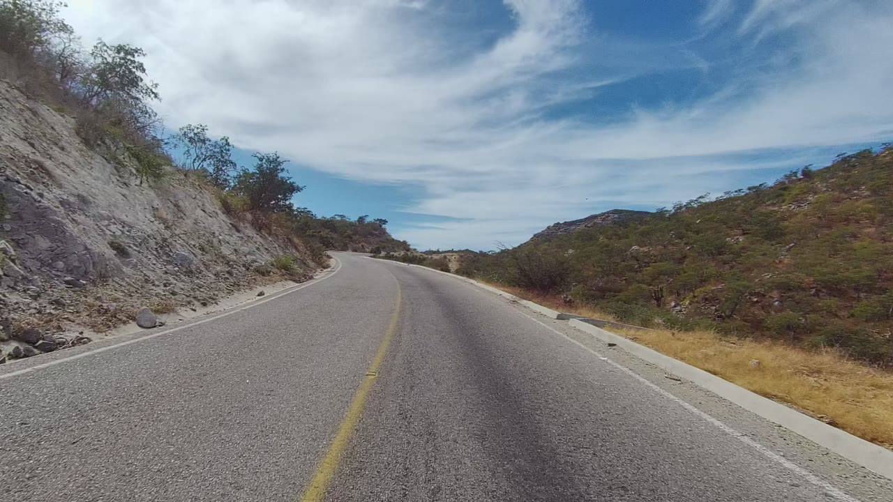 Road-level view in East Cape Los Cabos, Baja California Sur, showing a paved road bending left past rocky slopes, scrubby trees, dry grass, and no visible people.