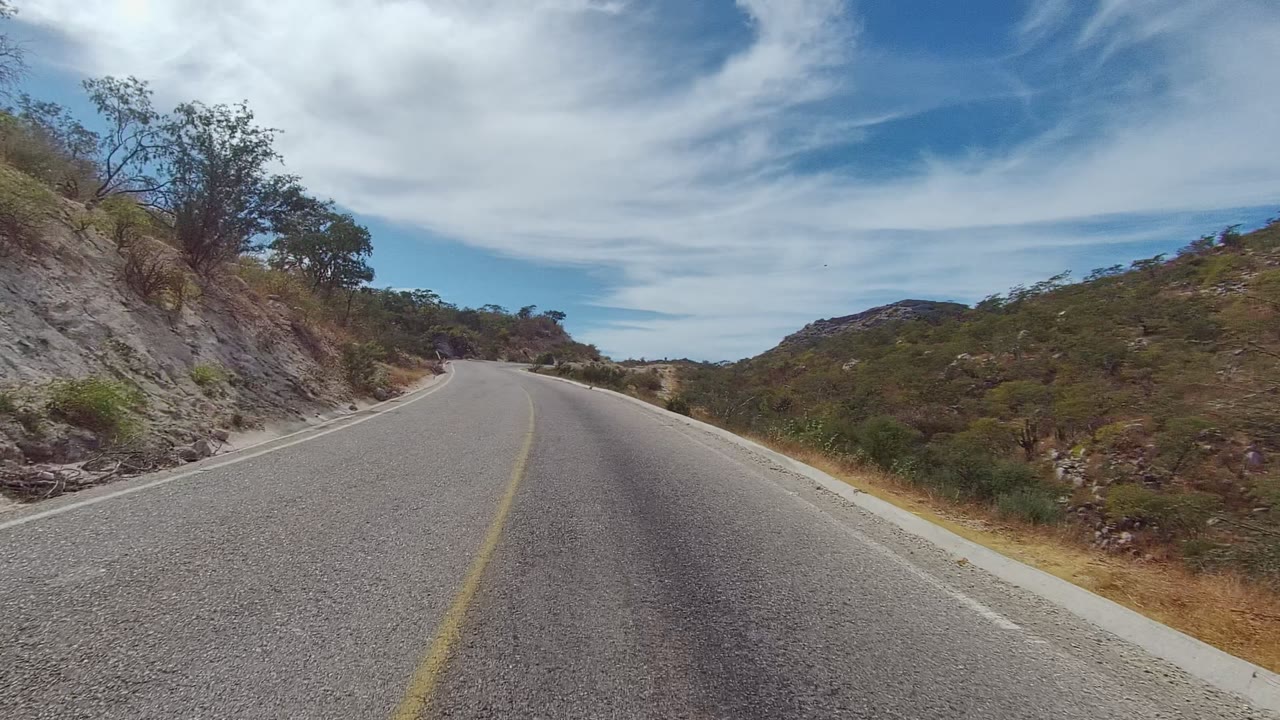 Forward road view with a yellow centerline, rocky hillside, shrubs, and clouded sky.