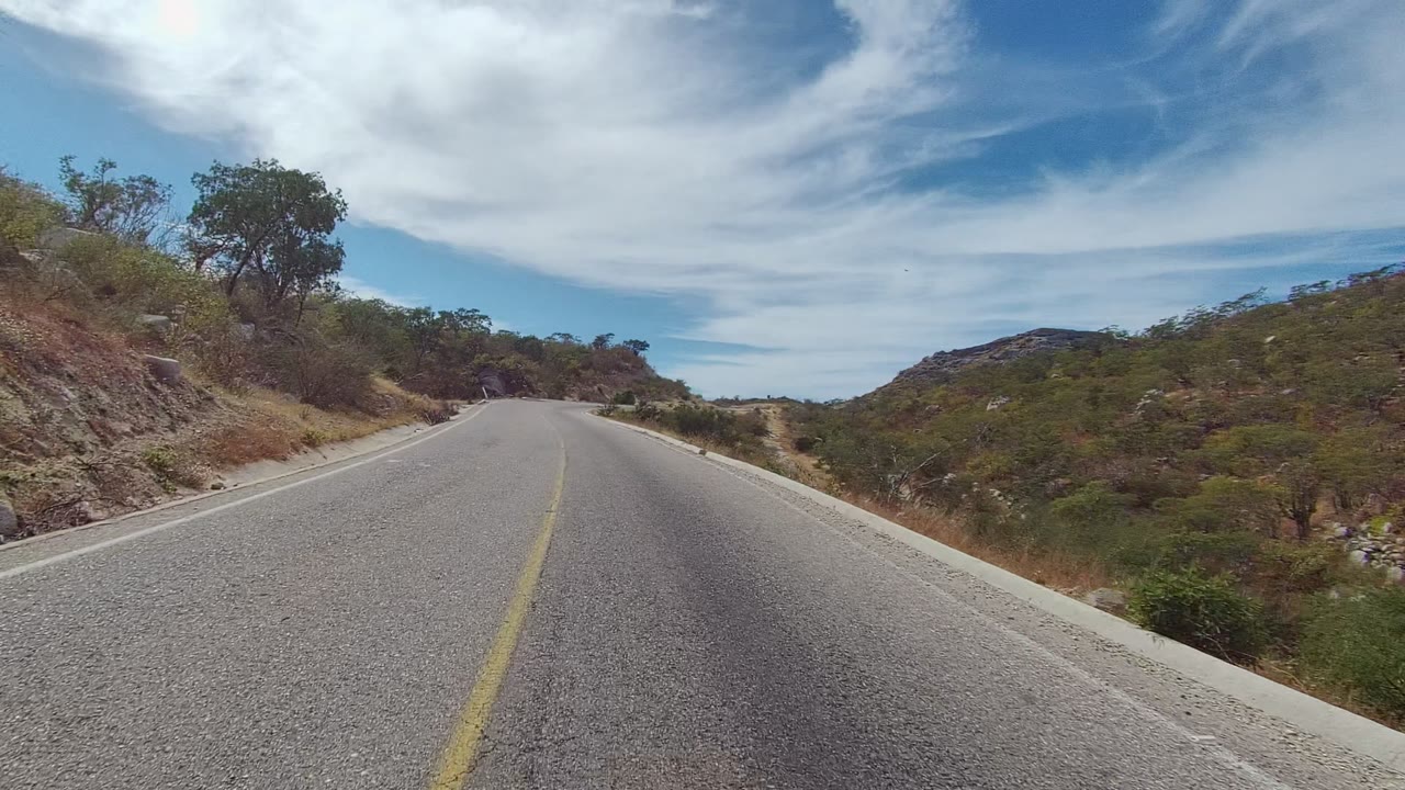 East Cape Los Cabos road frame showing the paved lane rising gently into a curve, with rocky slopes, dry roadside grass, scattered trees, and no visible people.