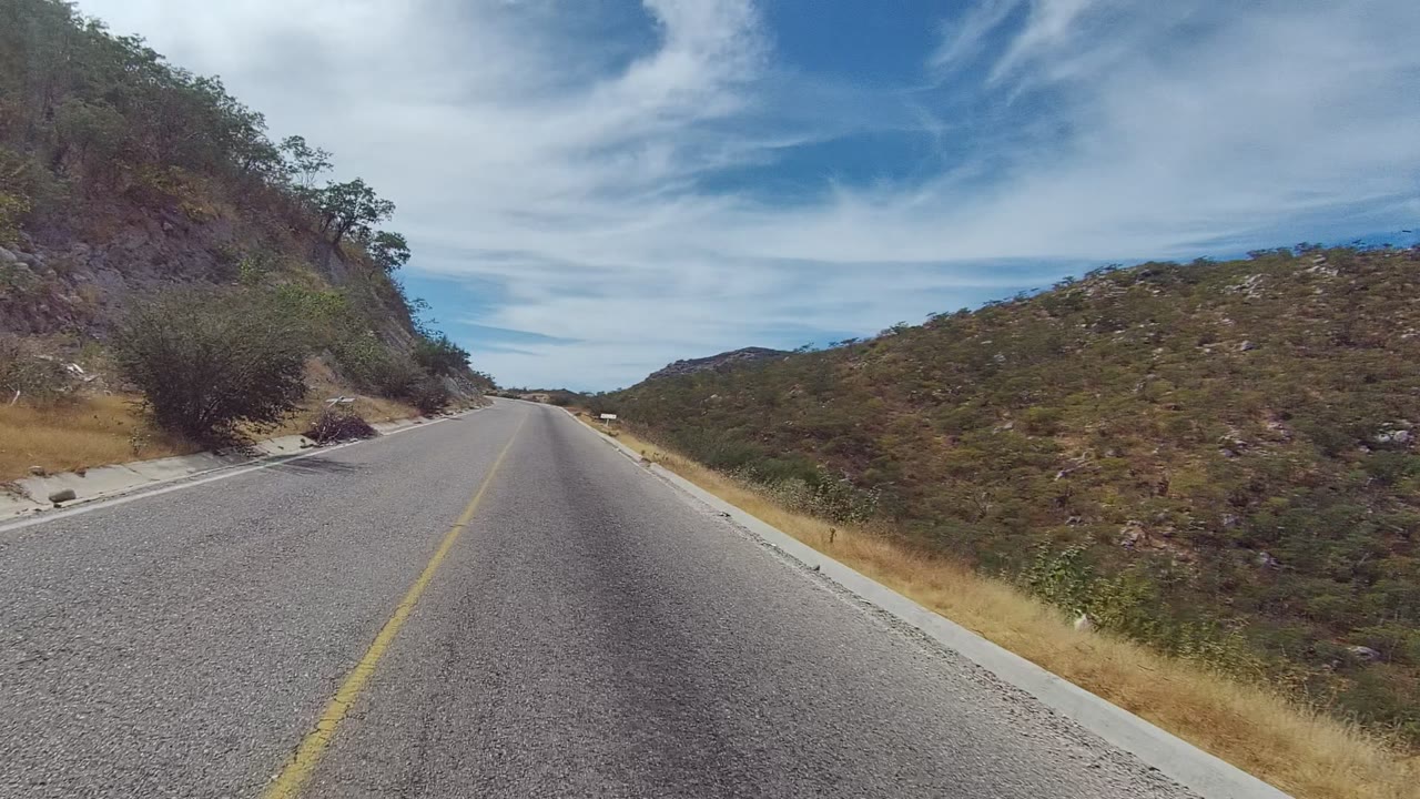 A paved road in East Cape Los Cabos runs between a close rocky hillside on the left and dry scrub-covered terrain on the right, with no visible people.