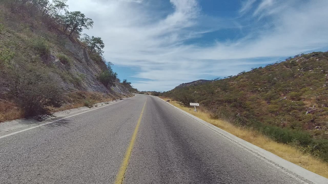 A straight stretch of paved road in East Cape Los Cabos passes dry grasses and rocky hills, with a small roadside sign on the right and no visible people.