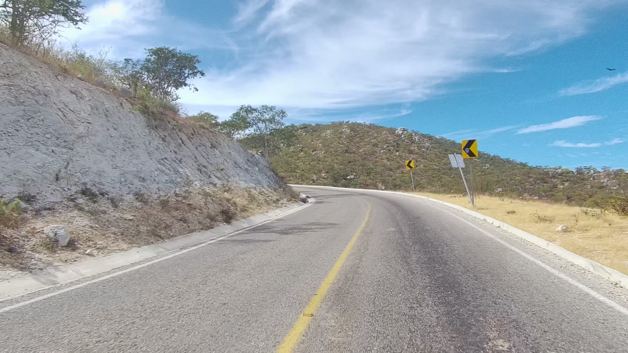 Opening frame of an East Cape Los Cabos road curving left beside a pale rocky cut, dry grass, scattered trees, blue sky, and no visible people.