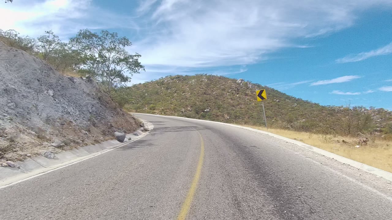 Winding road with a yellow curve sign and dry hillside scrub.