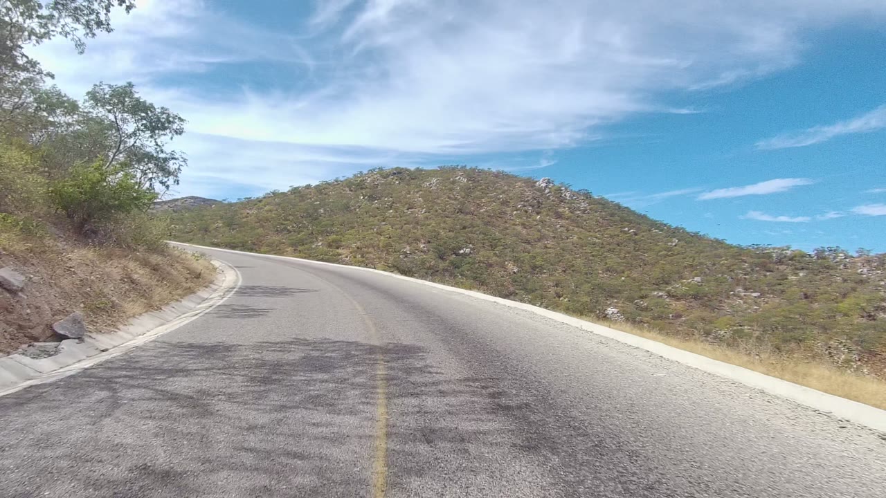 East Cape Los Cabos hillside road continuing through a left bend, with a broad scrub-covered hill ahead, pale shoulder edges, cloud streaks, and no visible people.