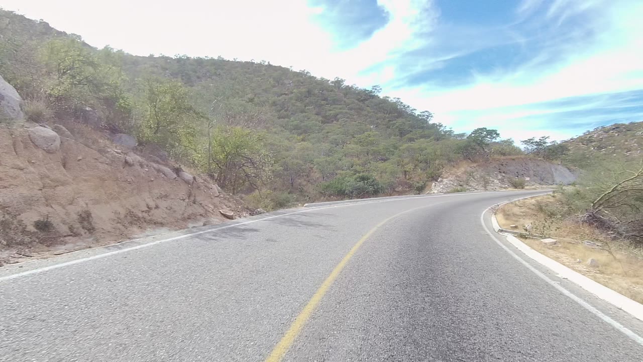 A paved road curves right through hills in East Cape Los Cabos, Baja California Sur, with a yellow center line, dry vegetation, rocky roadside, and no visible people.