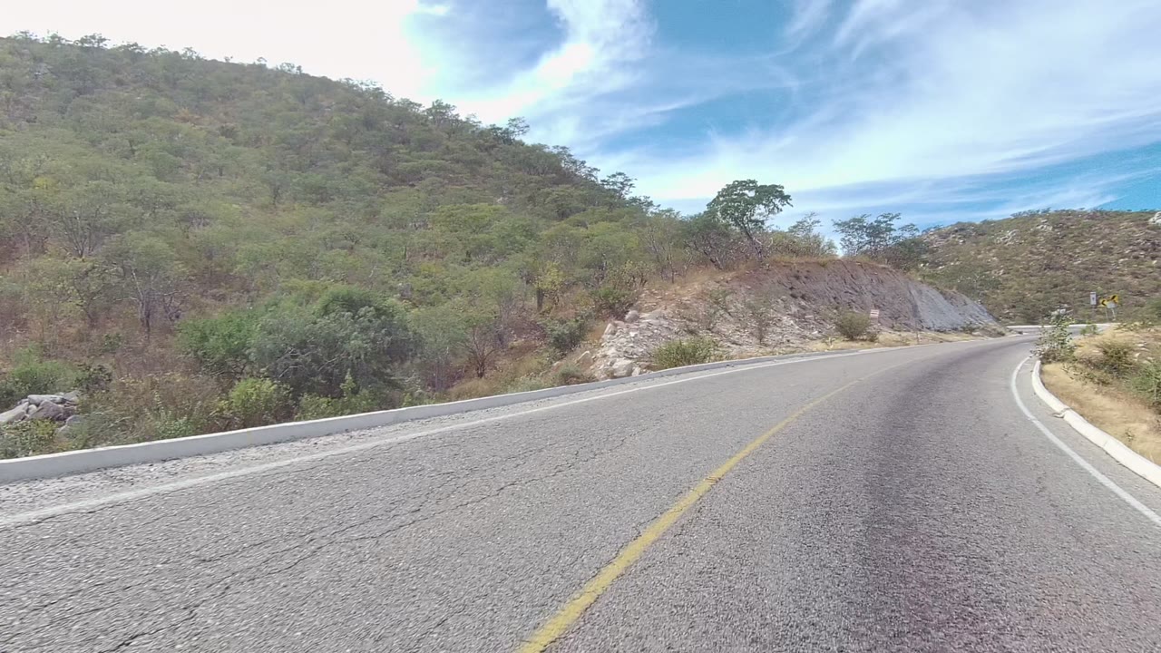 A winding road in East Cape Los Cabos, Baja California Sur, passes scrub and pale rock with curve warning signs ahead and no visible people.
