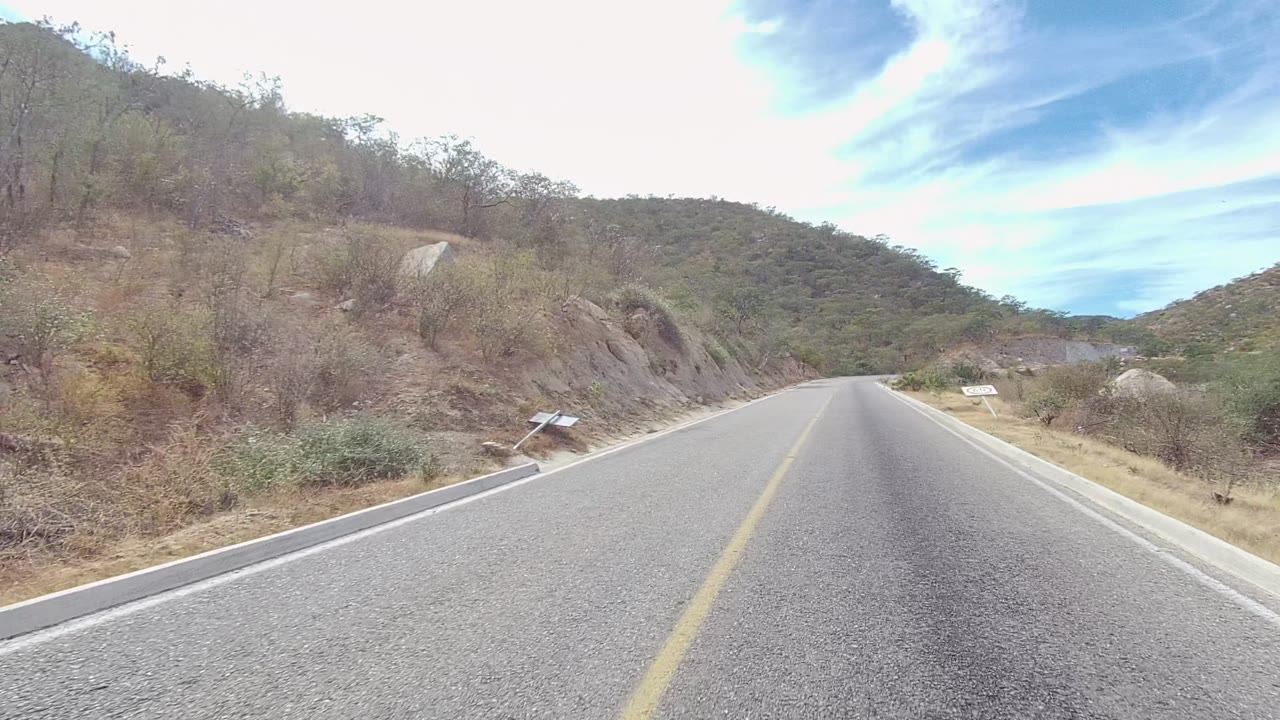 Road-level view in East Cape Los Cabos showing a paved two-lane road, yellow center line, dry scrub, rocky slopes, distant hills, and no visible people.