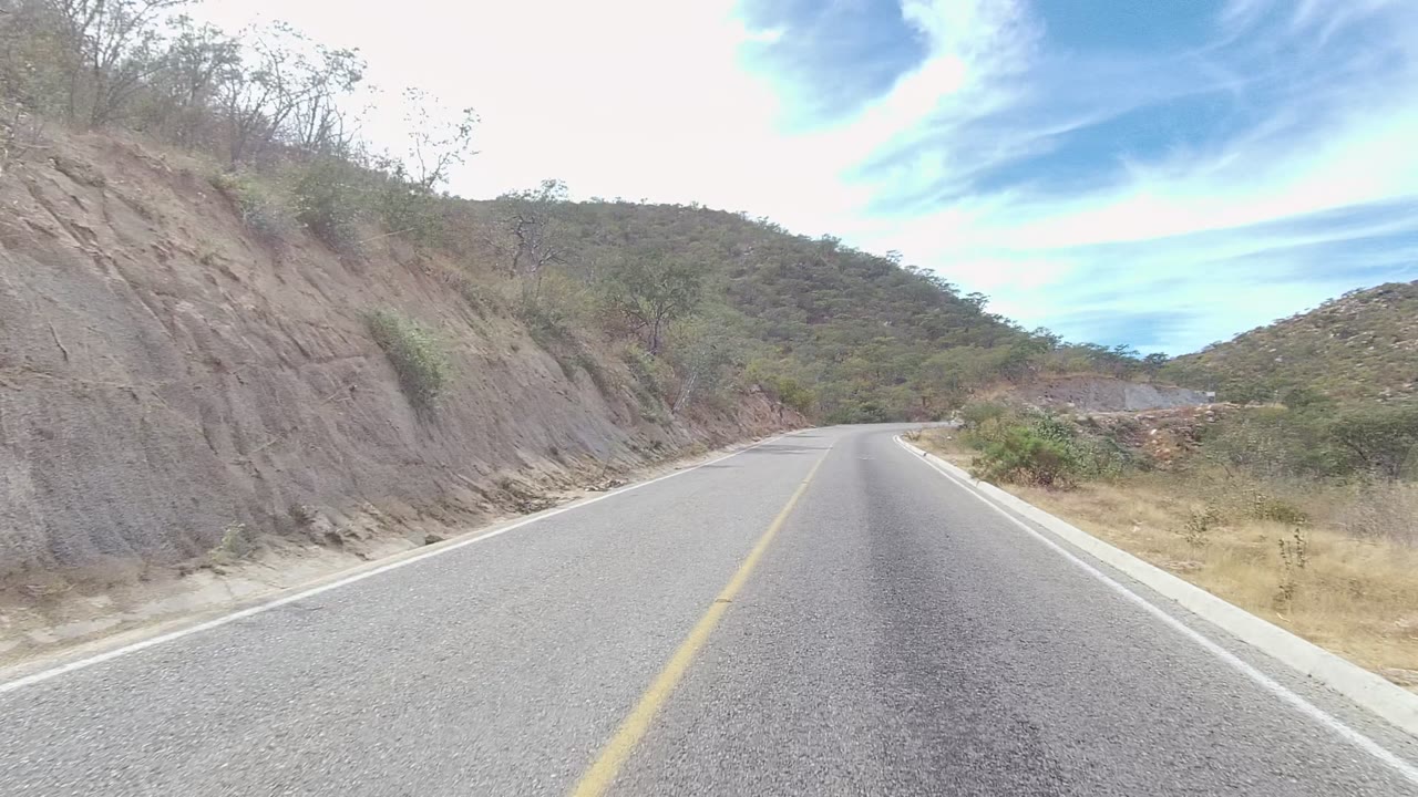 East Cape Los Cabos road view with the asphalt beginning to bend right, a steep rocky slope on the left, dry roadside plants, low hills ahead, and no visible people.