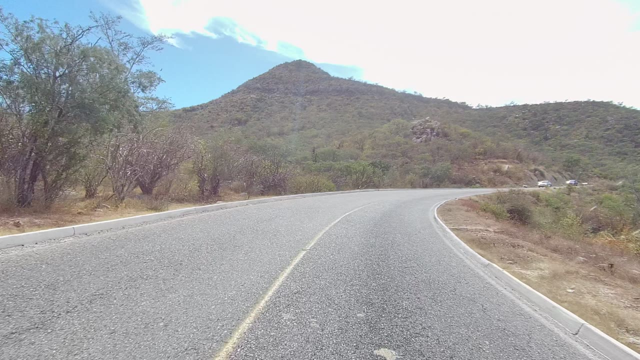 A curving asphalt road in East Cape Los Cabos, Baja California Sur, runs toward low green-brown hills under bright partly cloudy sky, with no visible people.