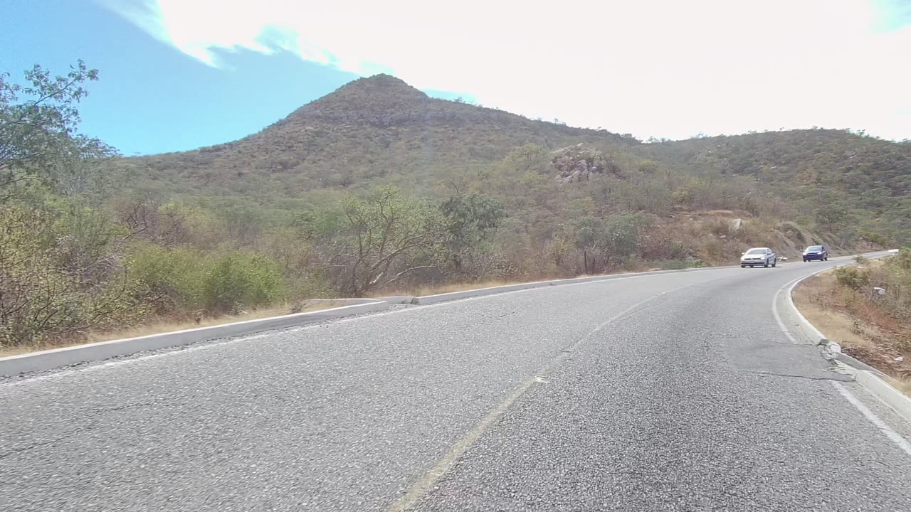 Two cars approach around a paved bend in East Cape Los Cabos, with scrub vegetation, rocky hillsides, and no visible people in the road scene.