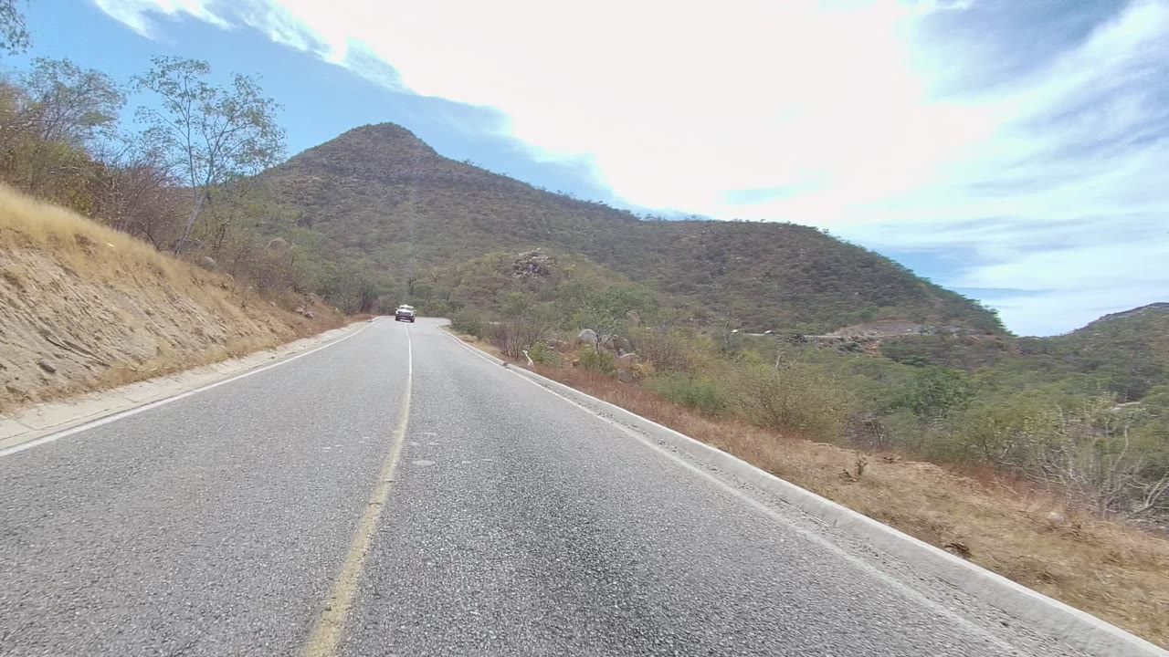 A distant car approaches on a paved East Cape Los Cabos mountain road bordered by dry grass, scrub trees, rocks, and low hills, with no visible people.