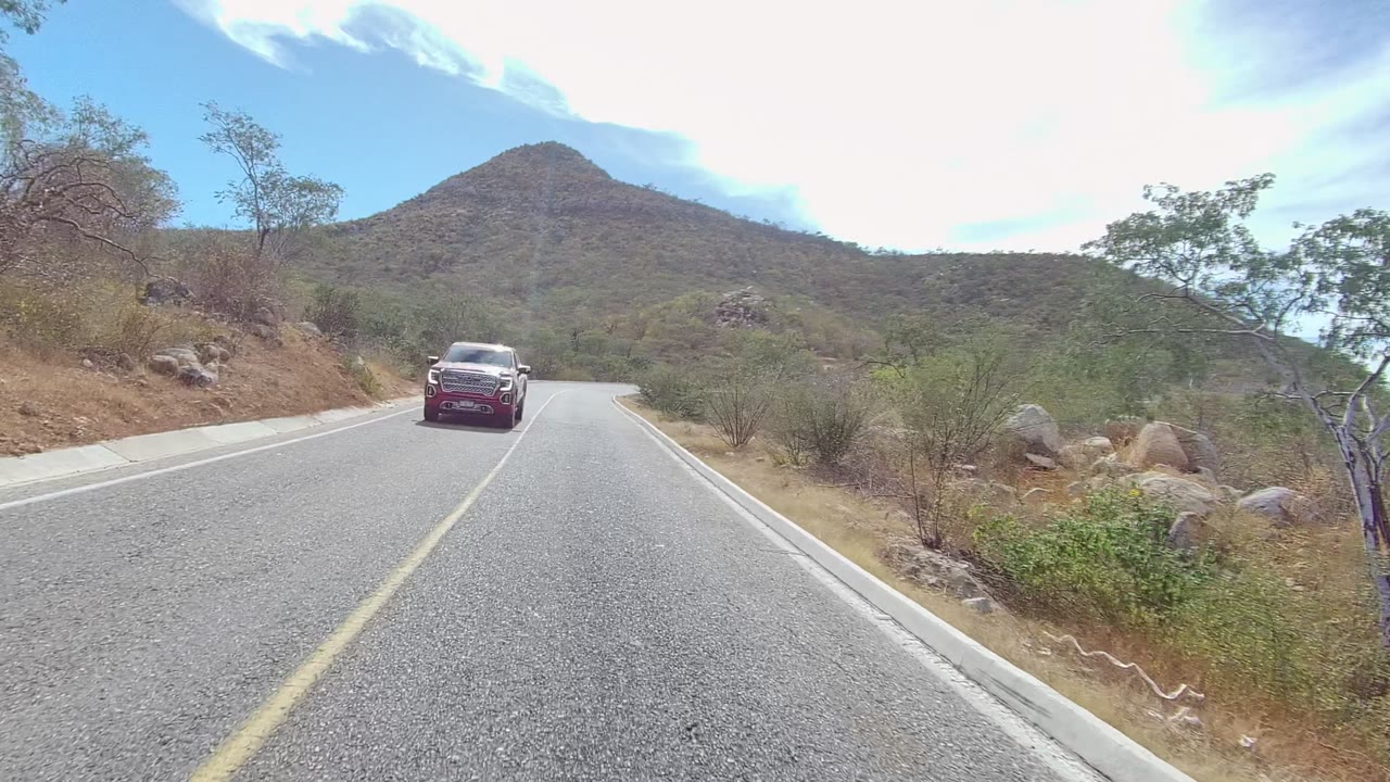 A car passes close to the camera on the East Cape Los Cabos road, with dry brush, rocks, a curving shoulder, green hills, and bright partly cloudy sky visible, with no visible people.