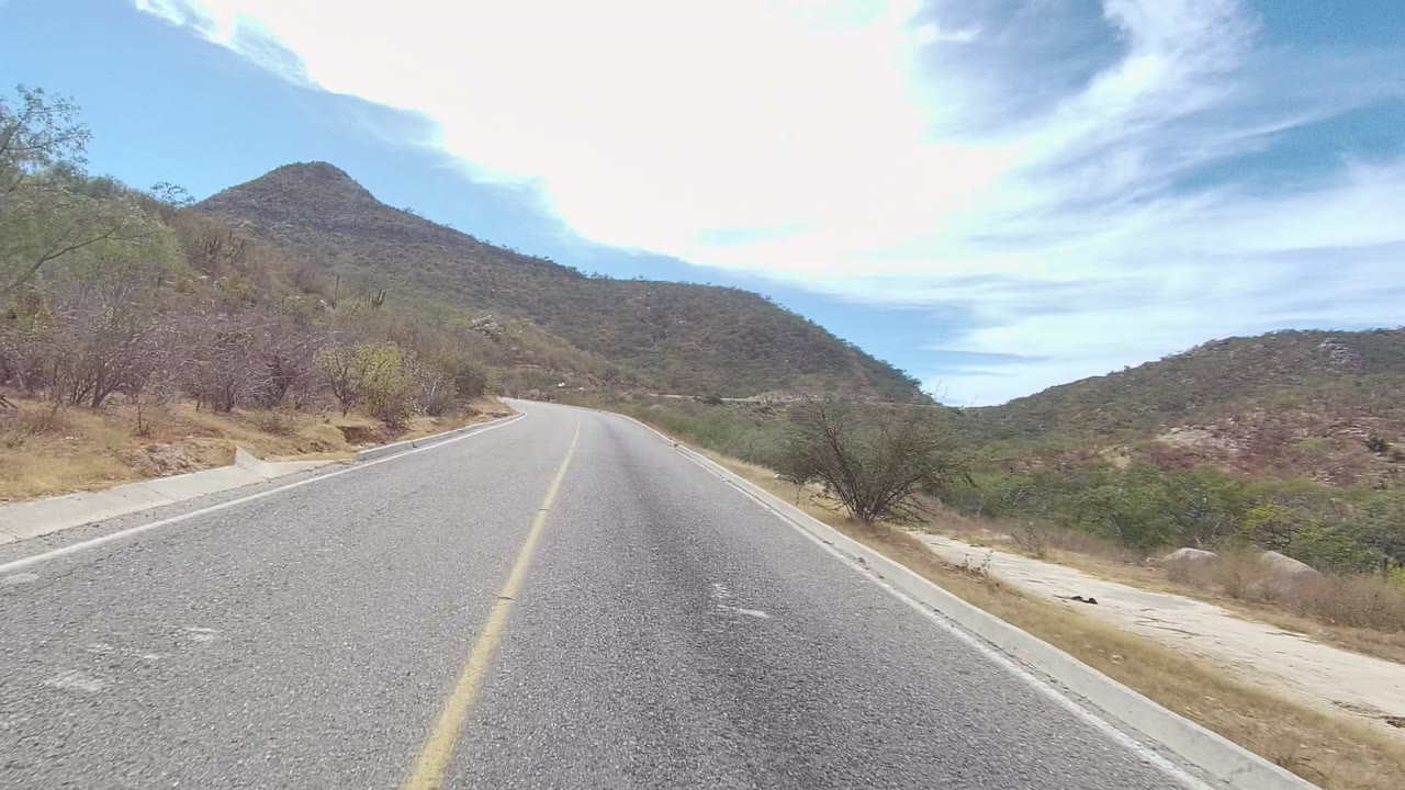 At 0:00, a paved road in East Cape Los Cabos curves left through arid hills with a yellow center line, sparse brush, low mountains, bright clouds, and no visible people.