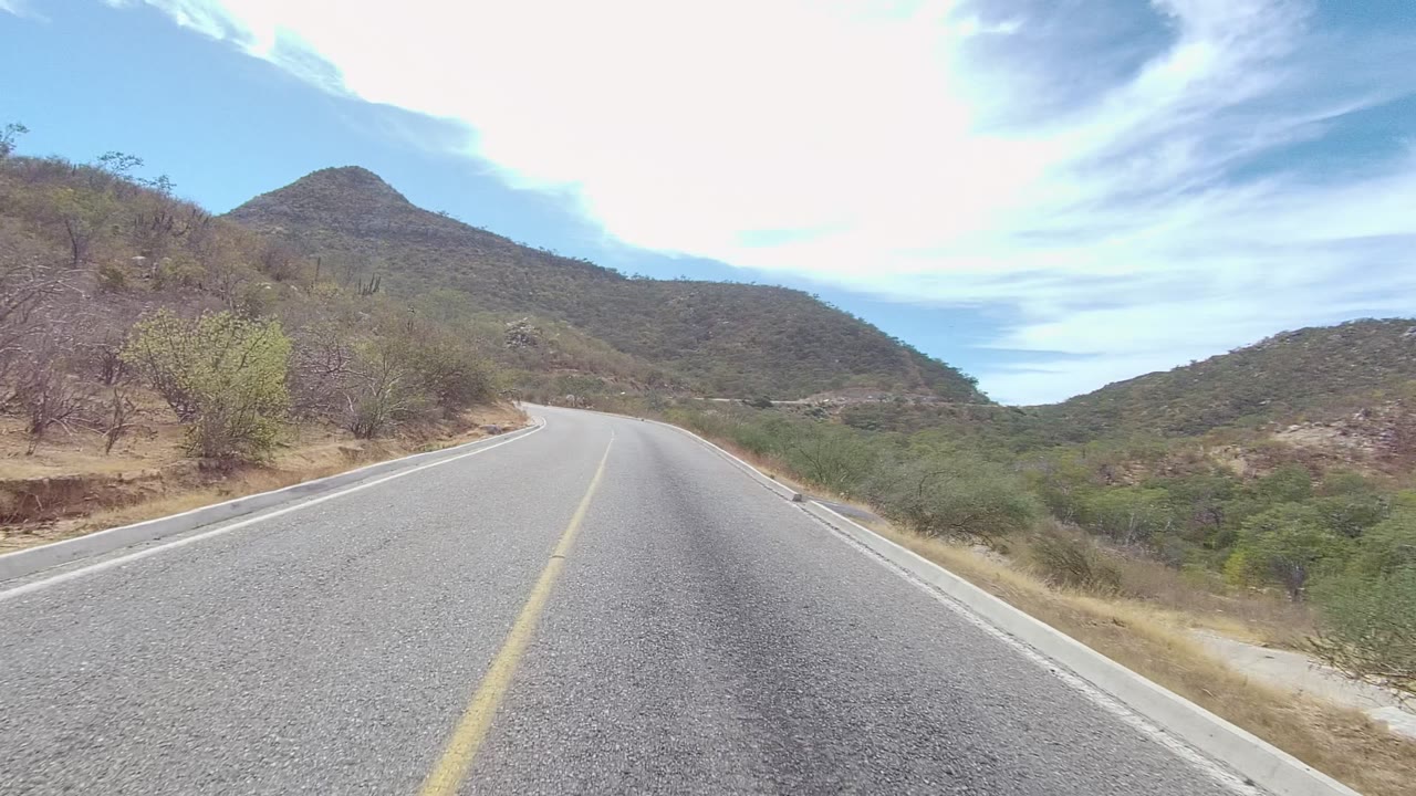 Two-lane road bending left beside scrub-covered hills.