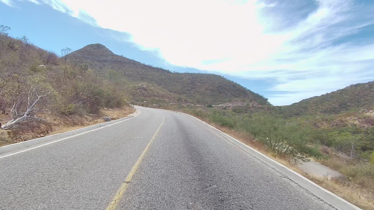 At 0:02, the road in East Cape Los Cabos curves around a dry slope with scrub trees on the left, green brush in the valley, distant hills, a bright sky, and no visible people.