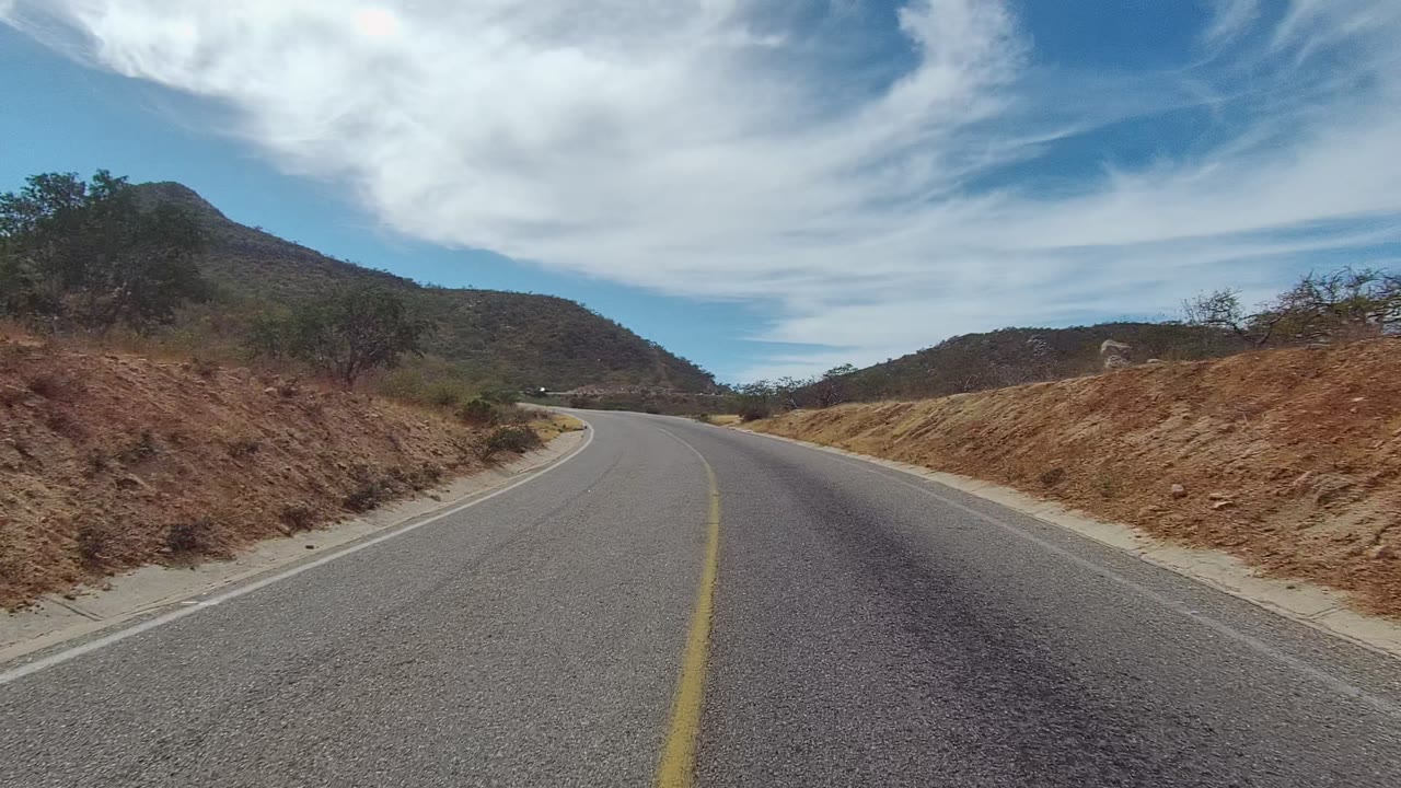 At 0:00, a paved rural road in East Cape Los Cabos curves between dry hills and sparse brush under a blue sky with white clouds, with no visible people.