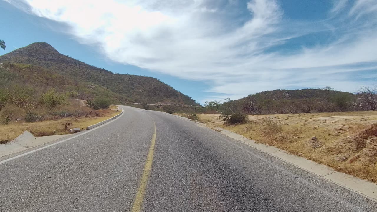 At 0:02, the empty paved road leads toward rounded dry hills with scattered desert vegetation and streaked white cloud cover in East Cape Los Cabos, with no visible people.