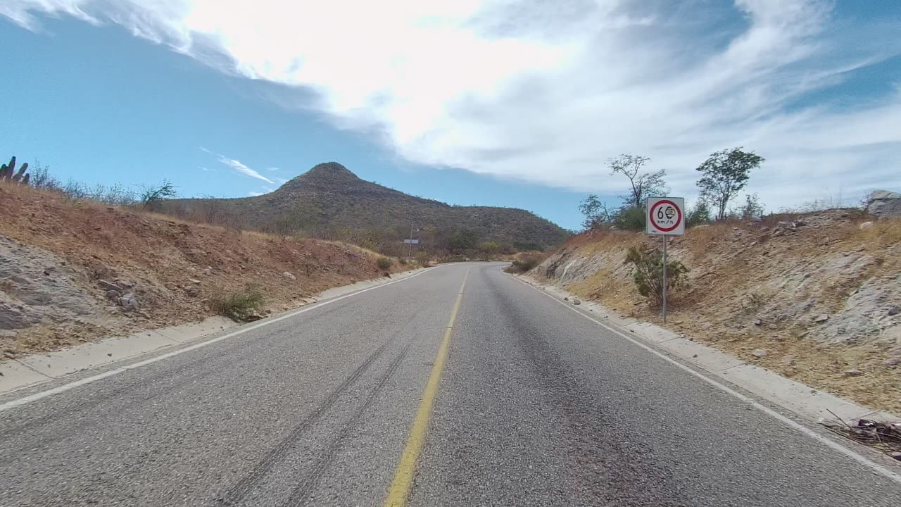 Road-level view in East Cape Los Cabos, Baja California Sur, with a two-lane road, roadside speed-limit sign, dry embankments, distant hill, bright sky, and no visible people.