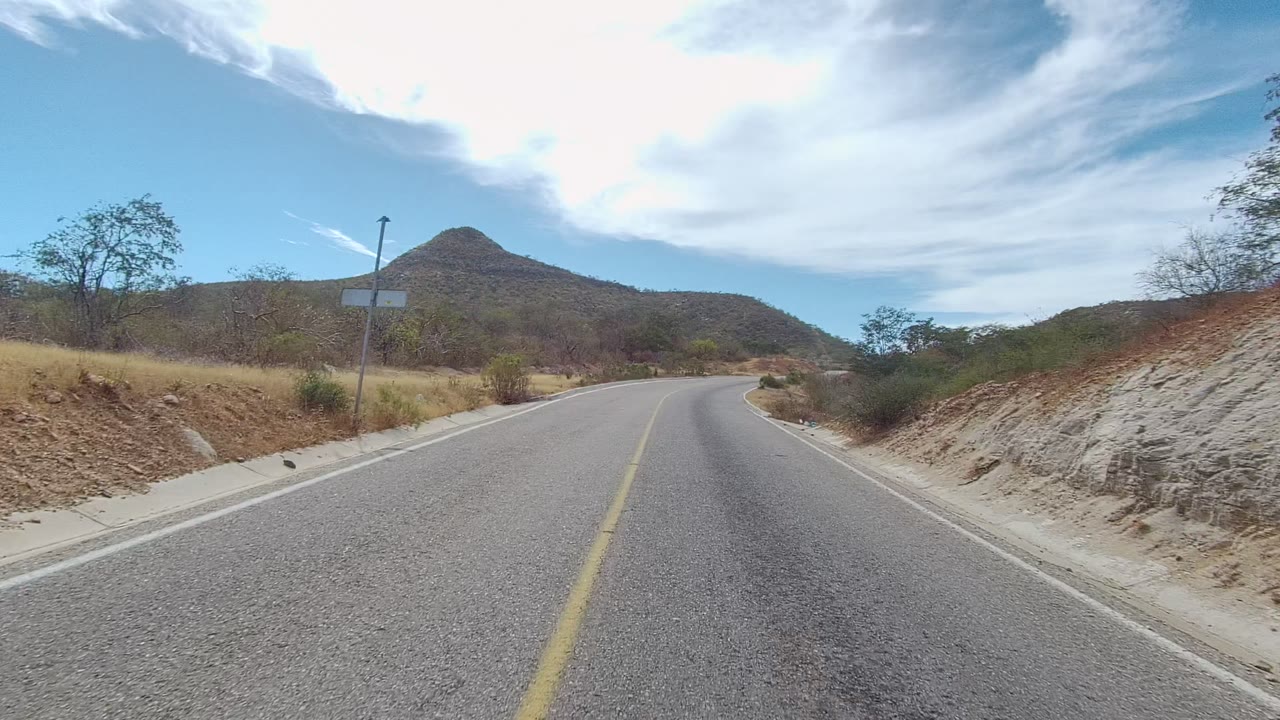 East Cape Los Cabos road scene with the two-lane pavement bending right, a roadside signpost, scrubby vegetation, dry slopes, distant hill, bright clouded sky, and no visible people.