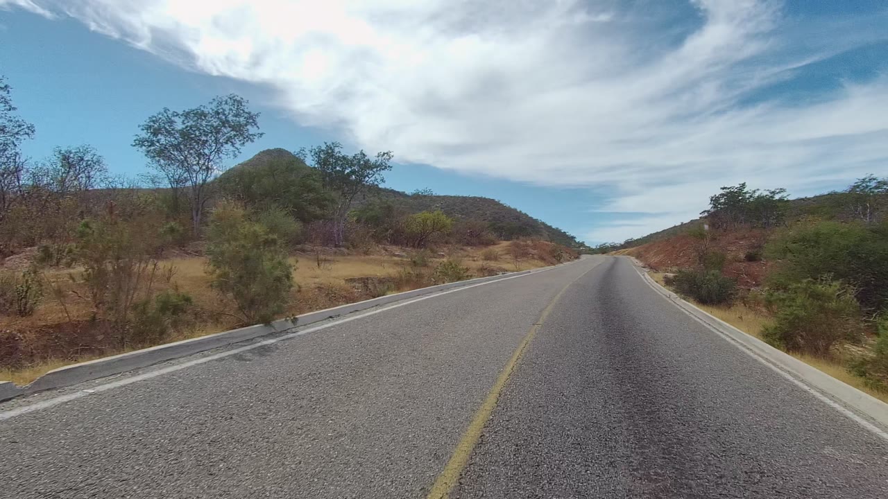 A paved two-lane road runs through dry hills and sparse shrubs in East Cape Los Cabos, Baja California Sur, with no visible people.