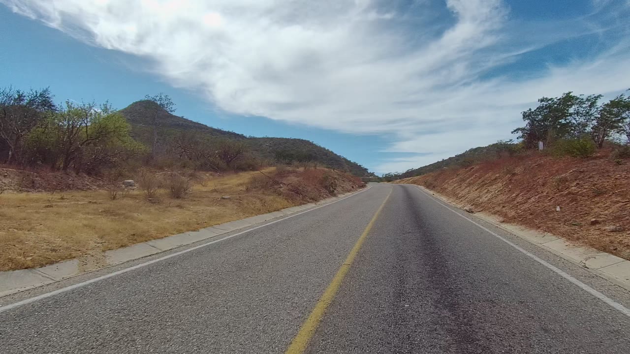 A slight road curve passes a red-brown cut bank and sparse roadside plants in East Cape Los Cabos, with a low green hill beyond and no visible people.
