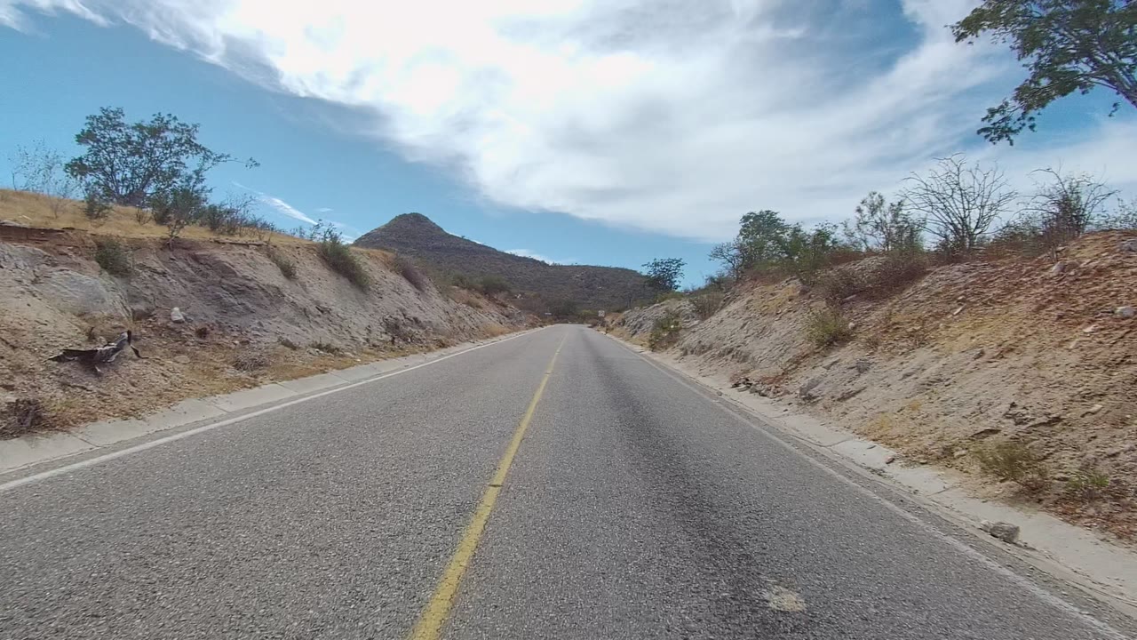 At 0:00, a paved road in East Cape Los Cabos runs between dry rocky hills with sparse shrubs, a distant rounded hill, bright clouds, and no visible people.