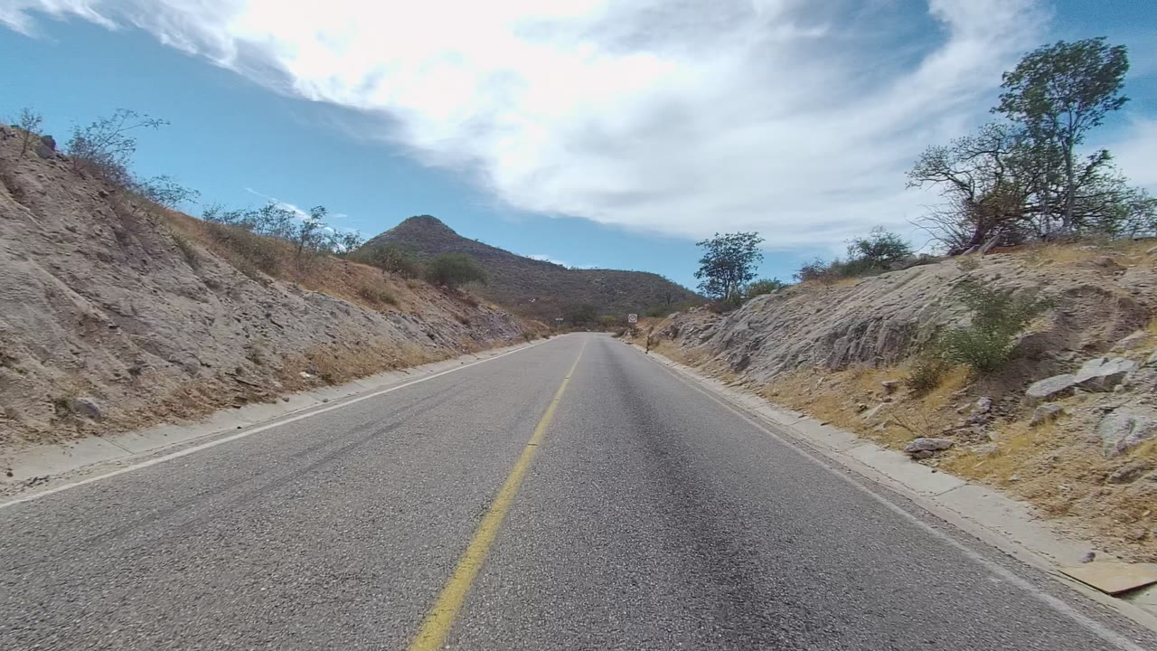 At 0:02, the paved East Cape Los Cabos road bends gently past rocky roadside slopes, dry brush, small trees, a rounded hill, and no visible people.