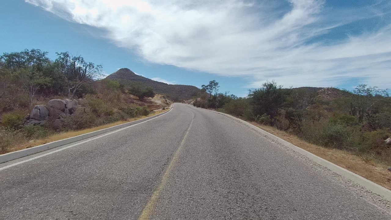 East Cape Los Cabos paved road centered between dry scrub hills and a rounded distant peak under bright white clouds, with no visible people.