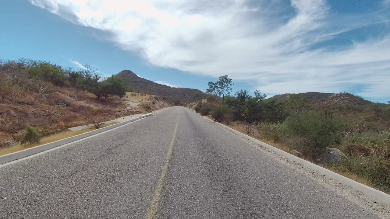 East Cape Los Cabos road scene with a wide asphalt foreground, pale curbs, scattered cactus and scrub on dry slopes, and a cloud band across the sky, with no visible people.