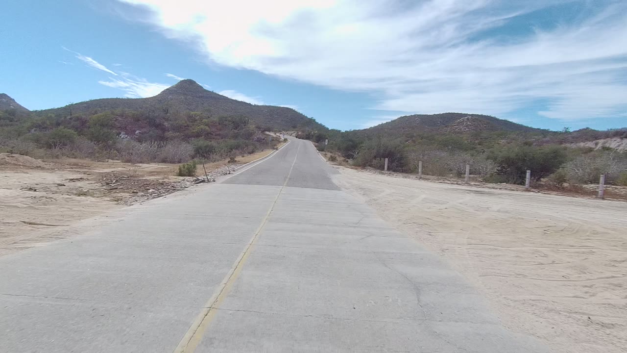 Start of the East Cape Los Cabos road clip showing a pale concrete road, sandy shoulders, sparse desert brush, distant hills, and no visible people.