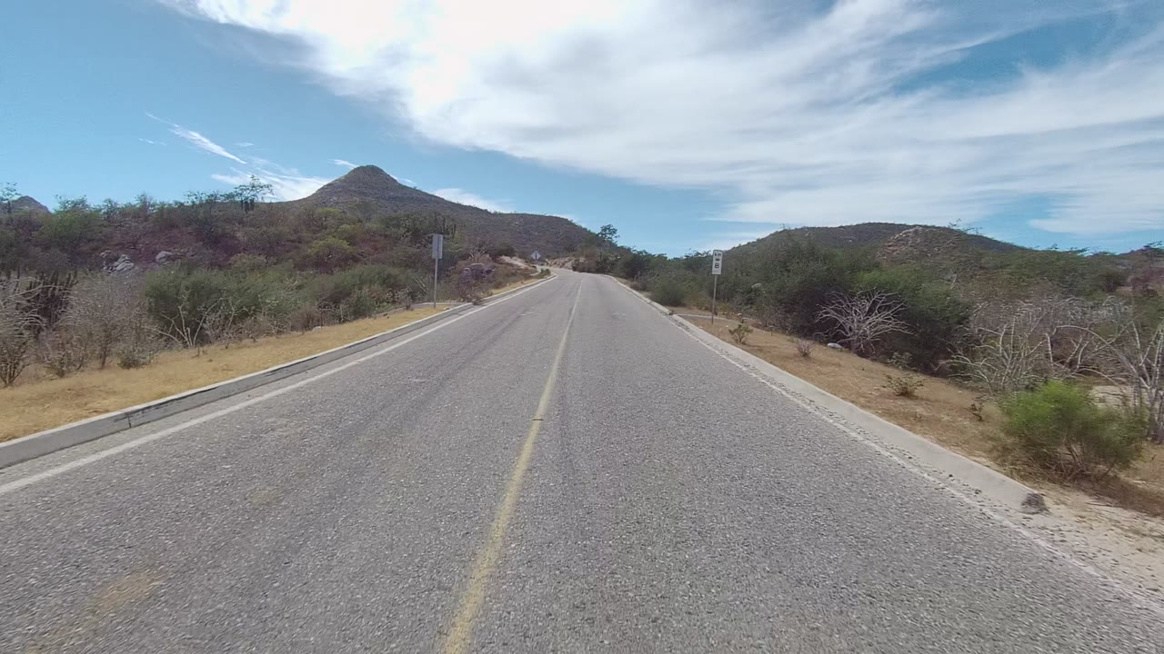 Centered view down an asphalt road in East Cape Los Cabos, with a faded yellow line, sandy edges, brush-covered hills, roadside signs, and no visible people.