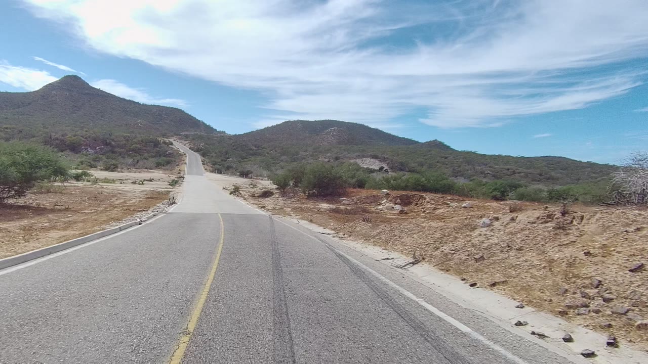 At the start of the East Cape Los Cabos clip, a paved road with a yellow center line leads toward low dry mountains and a pointed peak, with no visible people.