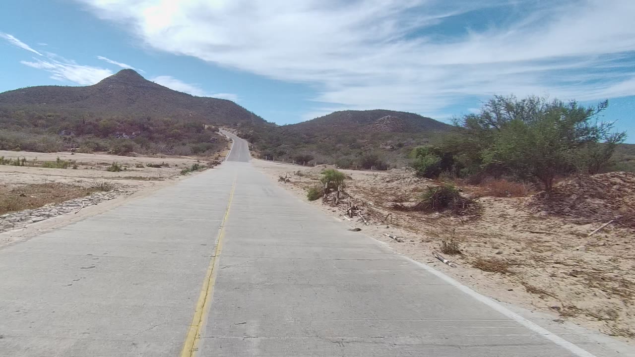 At 0:02 in East Cape Los Cabos, the centered concrete road continues toward low mountains, with sandy shoulders, sparse scrub, and no visible people.