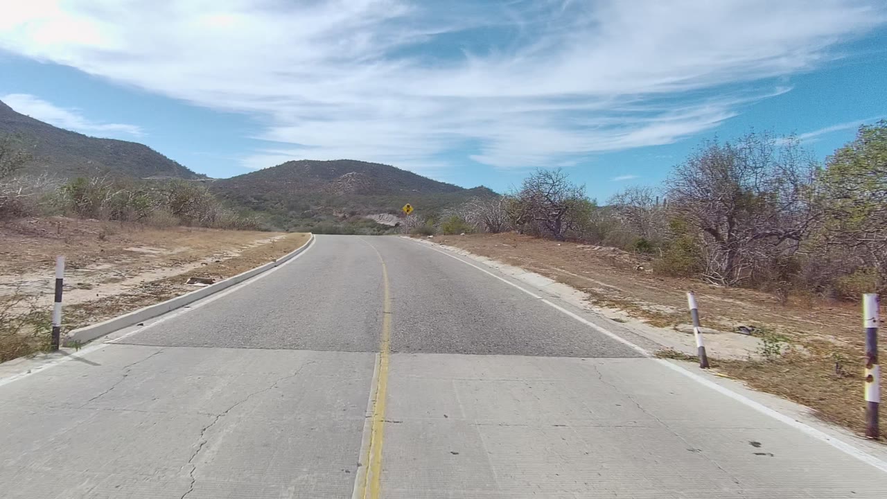 East Cape Los Cabos road passes over a small bridge, with black and white roadside markers, sparse scrub, mountains, and no visible people.