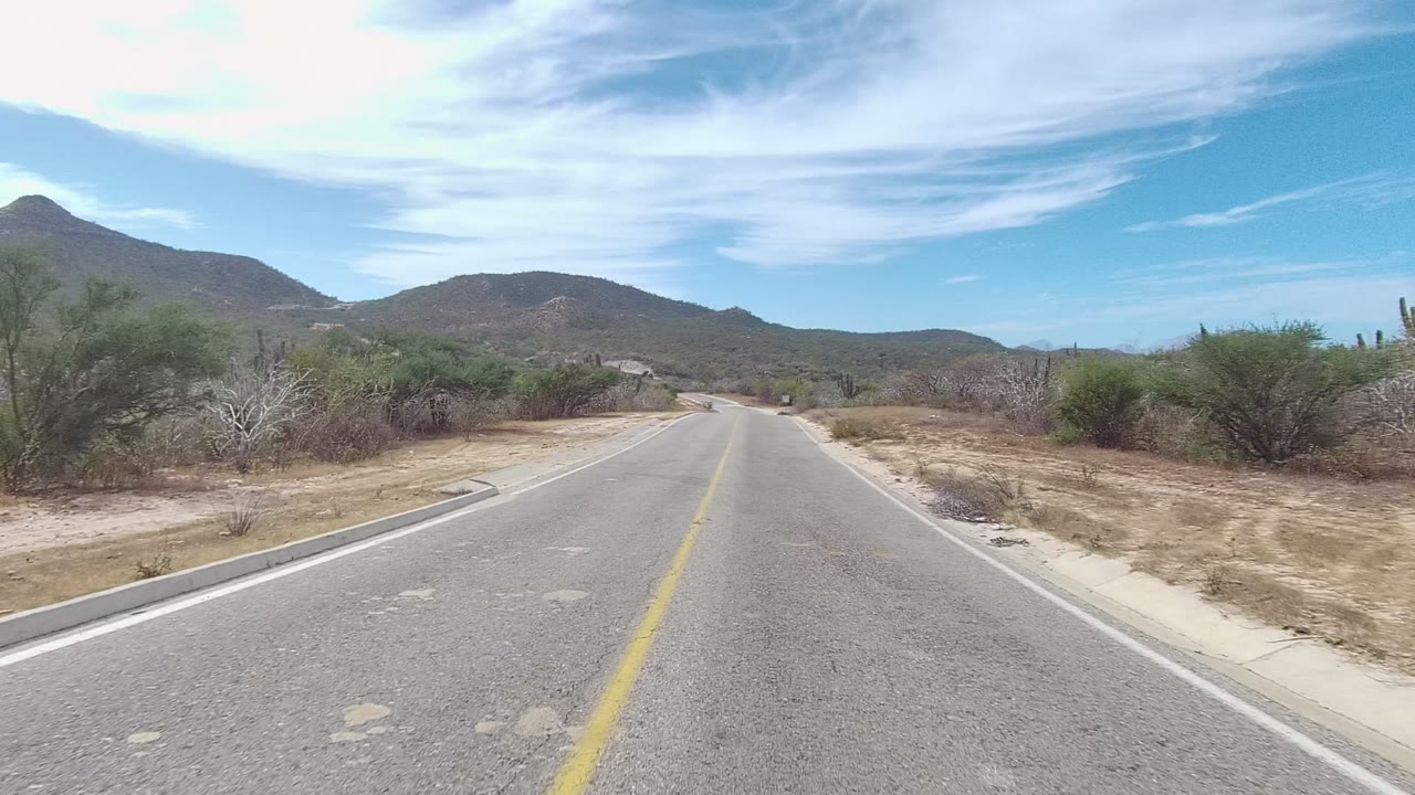 A paved East Cape Los Cabos road runs into dry desert terrain with low bushes, distant mountains, bright clouds, and no visible people.