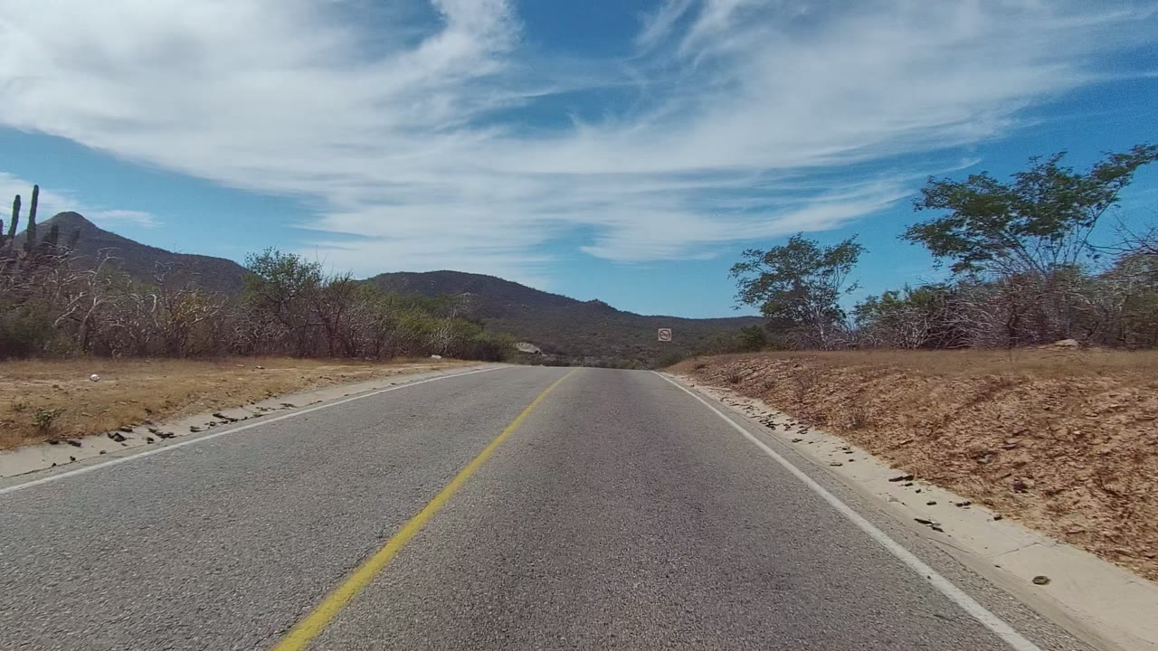 East Cape Los Cabos road view at the start of the clip, with a yellow center line, dry roadside brush, cactus at the left edge, distant mountains, broad white clouds, and no visible people.