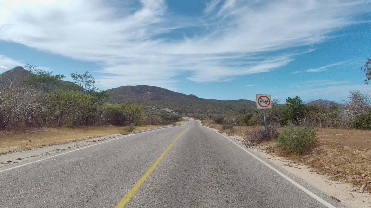 East Cape Los Cabos driving view with a no-passing-style roadside sign near the right shoulder, scrub and cactus lining the road, low mountains ahead, streaked clouds, and no visible people.