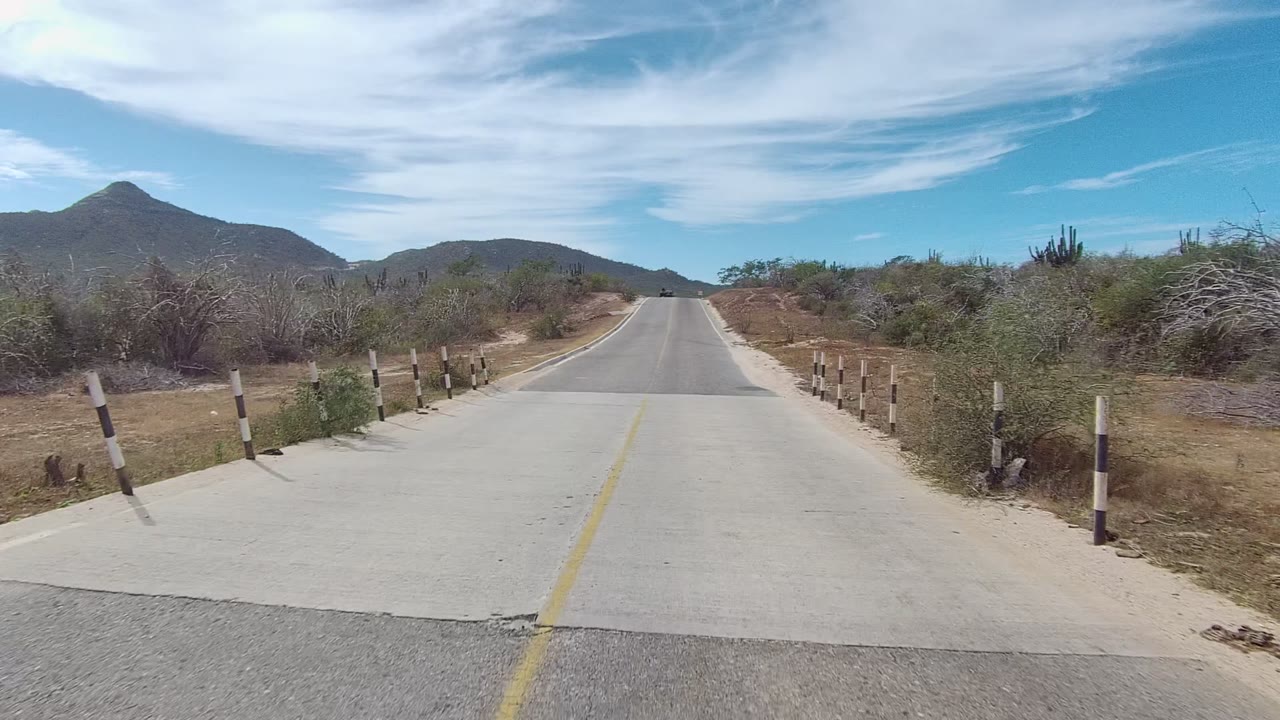 East Cape Los Cabos desert road with a yellow center line, roadside bollards, dry shrubs, distant mountains, bright clouds, and no visible people.