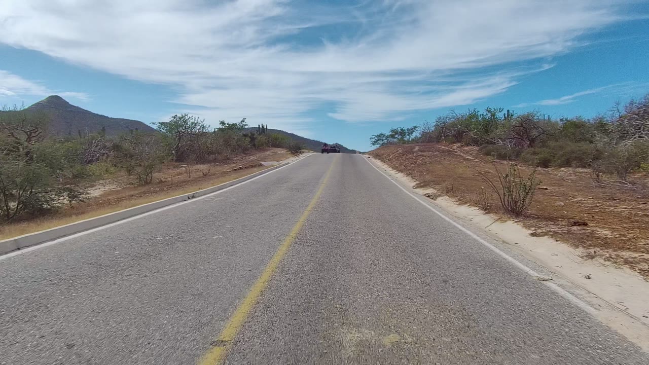 The dark SUV is closer on the East Cape Los Cabos asphalt road, framed by sandy shoulders, low desert vegetation, cacti, mountains, and no visible people.
