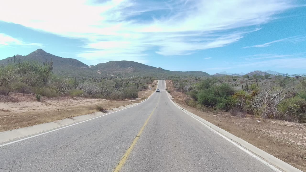 A straight paved road in Los Cabos, Baja California Sur, stretches through dry scrub and cactus toward low hills, with a small vehicle far ahead and no visible people.