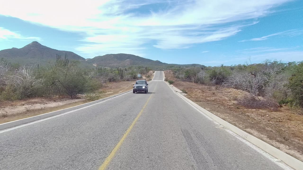 A dark vehicle passes close to the camera on the left side of the straight Los Cabos desert road, with cactus, scrub, hills, bright sky, and no visible people.