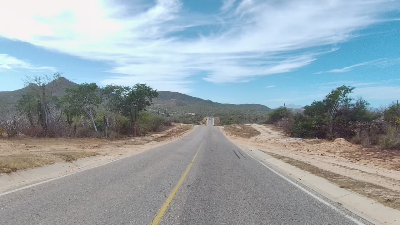 Start of the East Cape Los Cabos road view, with asphalt and a yellow center line leading through dry scrub toward distant hills, no visible people.