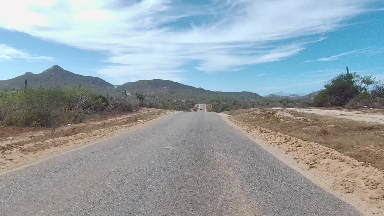 East Cape Los Cabos desert road view with a pointed mountain rising to the right of the road and low scrub lining the sandy shoulders, no visible people.