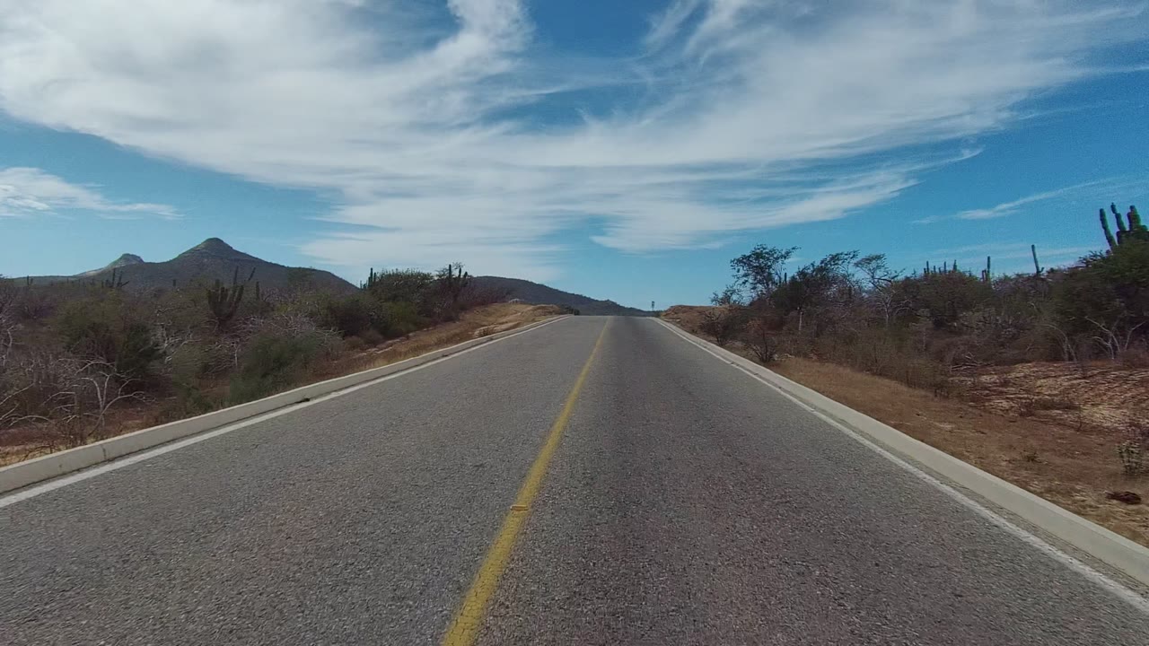 A paved road in East Cape Los Cabos, Mexico starts the clip with a yellow center line, dry roadside brush, cacti, distant hills, partly cloudy blue sky, and no visible people.