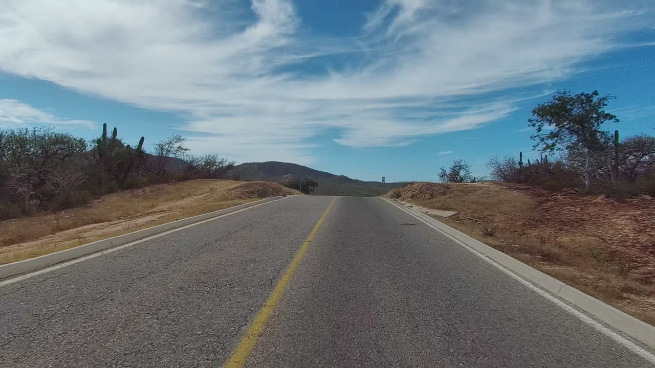 The road view in East Cape Los Cabos, Mexico shifts slightly right, showing a paved curve bordered by dry earth, bushes, cacti, distant mountains, blue sky, and no visible people.