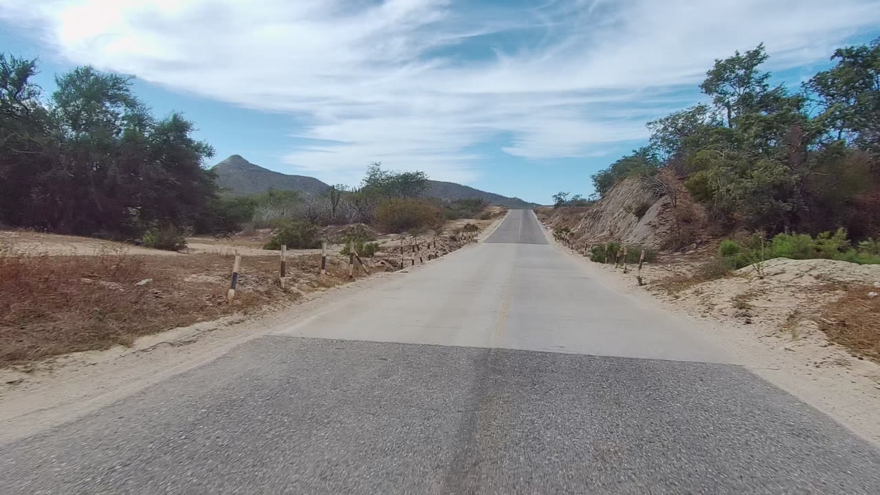 A paved road in East Cape Los Cabos, Mexico runs toward low hills, with sandy shoulders, sparse brush, blue sky, streaked clouds, and no visible people.