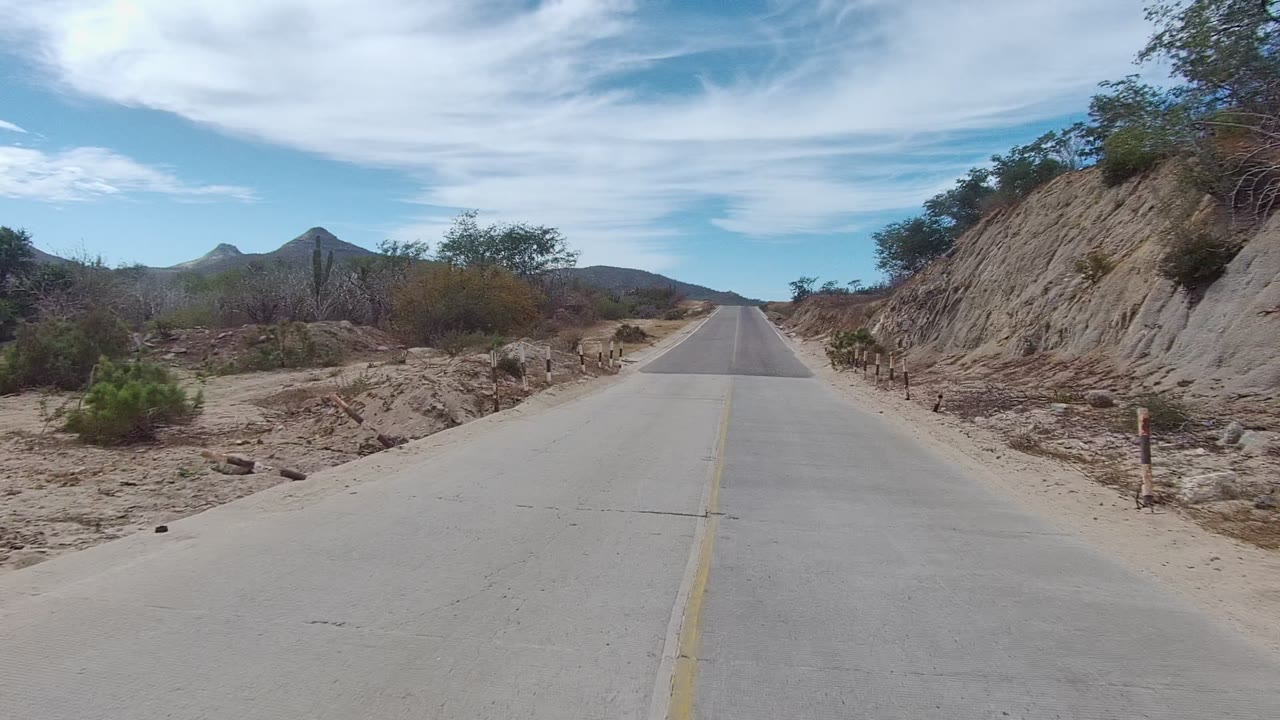 Road beside a rocky dirt cut with scrub and hills ahead.